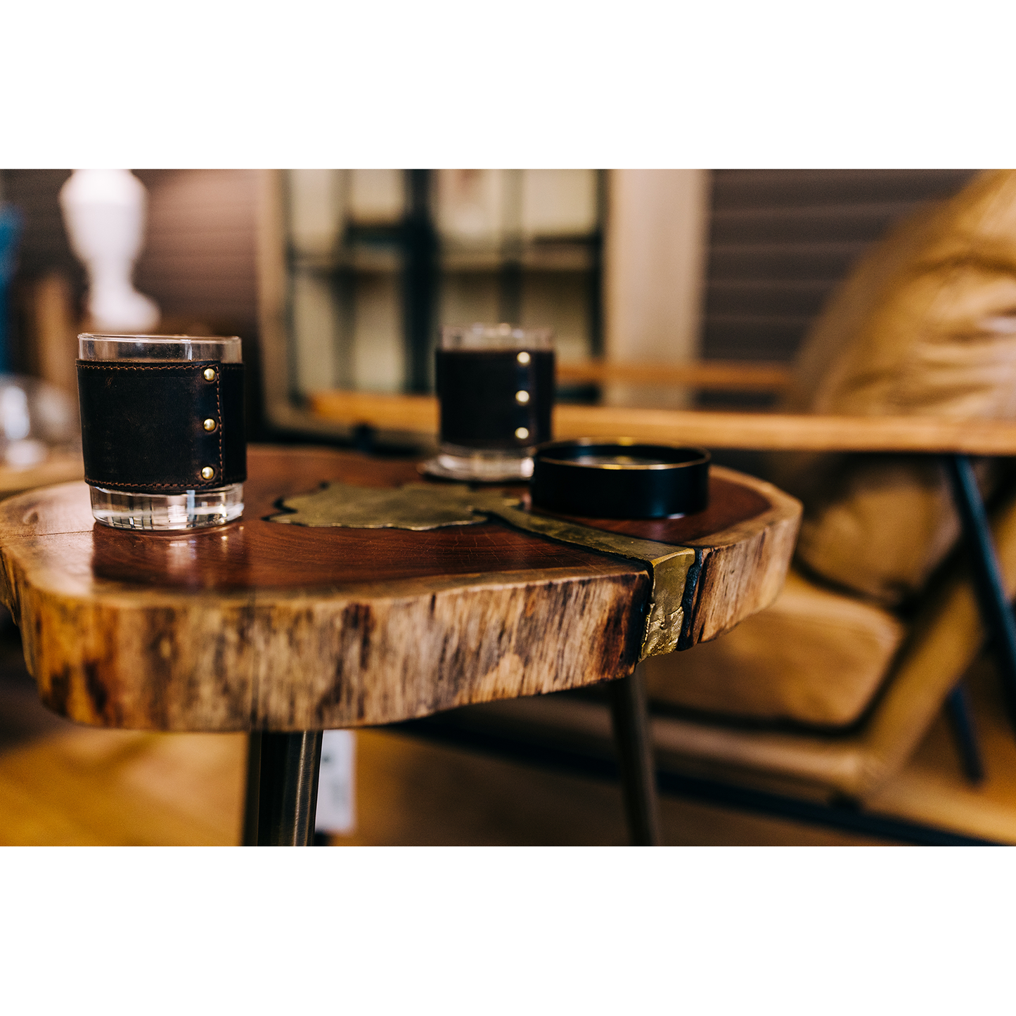 Close-up of the Brass Inlay Side Table crafted from rustic acacia wood. Two small glasses filled with dark liquid rest next to a black dish on the table, while a leather chair and softly lit interior in the background create a cozy, warm atmosphere.