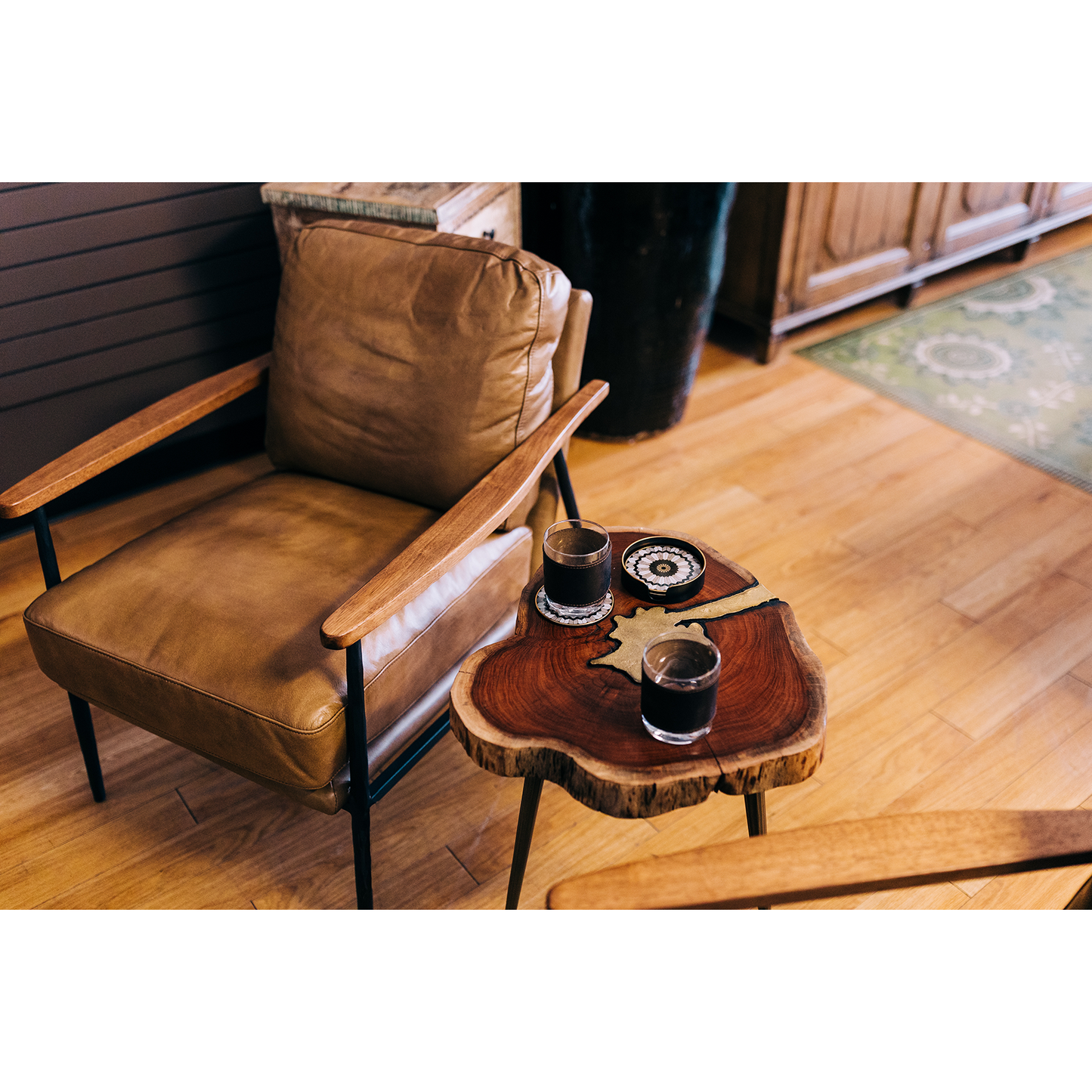 Cozy seating area with a tan leather armchair and the Brass Inlay Side Table made of rustic acacia wood. The table holds two small glasses filled with a dark liquid and three decorative coasters. The space has warm wooden flooring and a partially visible patterned rug in muted green tones.