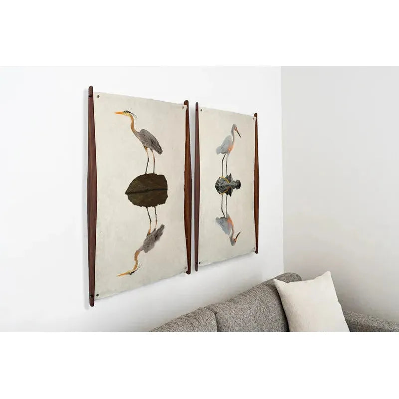 Two framed heron prints with water reflections hang above a gray sofa on a white wall. Frameless Wood/Steel Hanger, Curved ensures secure, damage-free photography display in this modern living room.