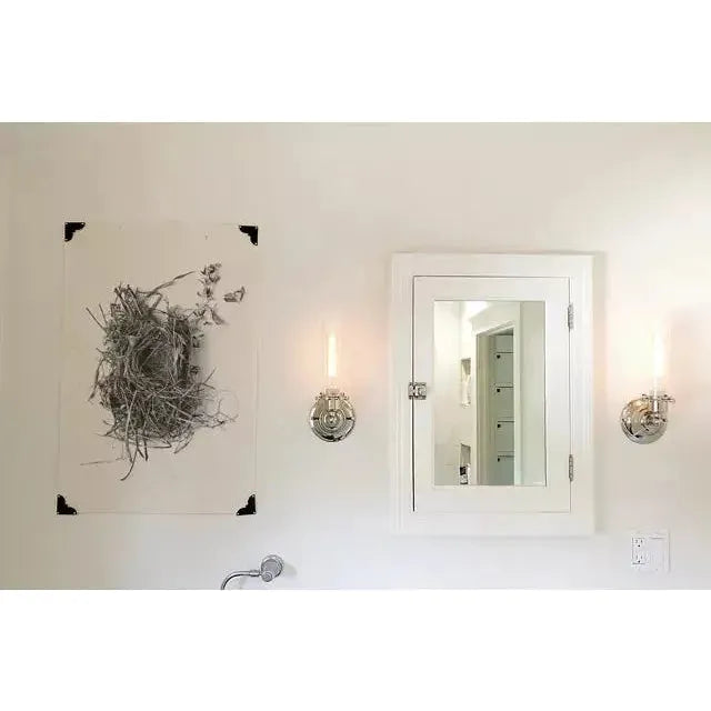 A bathroom wall features a white cabinet with a mirrored door at the center, flanked by two modern, cylindrical wall sconces with exposed bulbs. To the left of the cabinet is a framed black-and-white photograph hung using Corner Hangers. The background is a clean, white wall.