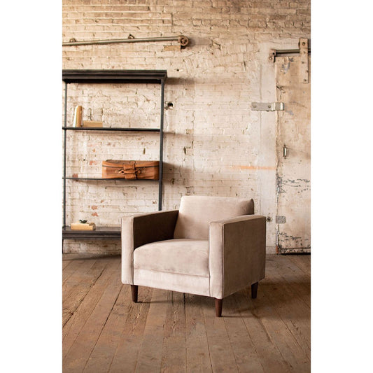 A contemporary polyester velvet Valerie Accent Chair in cobblestone color is positioned on a worn wooden floor against an exposed brick wall. To the left, a metal and wood shelf holds books and a leather bag, evoking a warm, rustic ambiance.