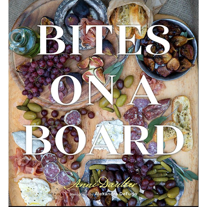 A charcuterie board featuring an array of foods such as clusters of red grapes, slices of figs, an assortment of cured meats and cheeses, olives, pickles, and crusty bread. The overlaid text says "Bites on a Board" with contributions from author Anni Daulter and photographer Alexandra DeFurio.
