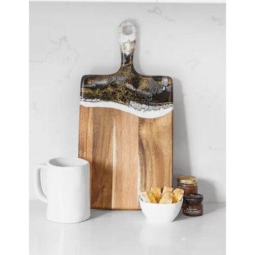 The Onyx Cheese Board, made of acacia wood with a black, white, and gold eco-friendly epoxy resin handle, stands upright. In front are a white mug, a bowl of chips, and two small jam jars on a white countertop against a light background.