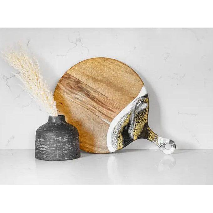 The Onyx Cheese Board, a round Acacia board with a handle and eco-friendly white and gold epoxy accent, leans against a white marble wall next to a black ceramic vase filled with dried pampas grass on a sleek white surface.