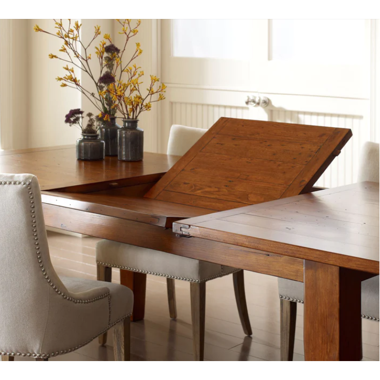 An Extension Dining Table, Large (72"/96"), crafted from recycled wood, with an extendable leaf partially lifted to showcase the extension mechanism. The table is surrounded by beige upholstered chairs with stud detailing. A centerpiece with branches in bottles graces one end of the table as light streams in from a window in the background.