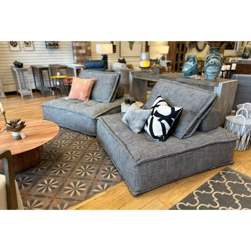 A furniture showroom displays two modern gray Prime Princeton Sofa Loungers with decorative cushions, a small wooden coffee table, and assorted decor items on shelves behind. The floor features a patterned rug.
