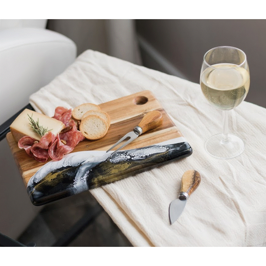 The Onyx Cheese Board is topped with cheese, sliced salami, rosemary, bread slices, and a cheese knife on a white cloth next to a glass of white wine. Another knife rests nearby—perfect for entertaining guests in style.