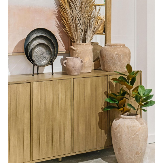 A wooden sideboard with decorative metal plates, textured clay vases, and dried pampas grass stands beside a large terracotta pot holding a green plant. Neutral tones and natural textures accentuate the gold rim of the Leonard Tray in front of an abstract artwork.