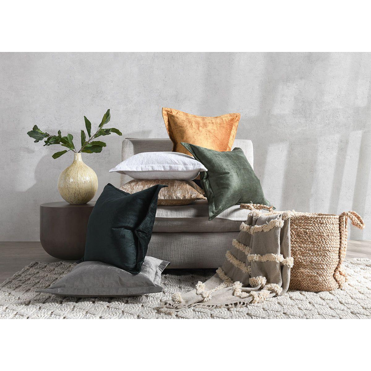 A cozy living room setup features a grey bench against a textured grey wall, adorned with a beige vase holding green branches. Assorted Lapis Throw Pillows in multiple colors, including green, orange, white, and grey, are arranged on the bench and floor. A woven basket filled with a beige tassel throw sits on a cream rug.