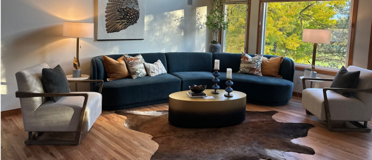 furniture for sale. a Rustic living room with a rough-hewn wooden table behind beige armchairs. Table holds white flower sculptures on golden stands. Black sofa with pink cushions and bookshelves in the background.