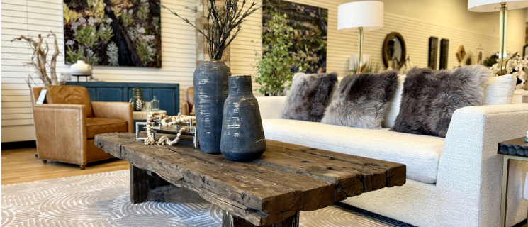 furniture for sale. a Rustic living room with a rough-hewn wooden table behind beige armchairs. Table holds white flower sculptures on golden stands. Black sofa with pink cushions and bookshelves in the background.