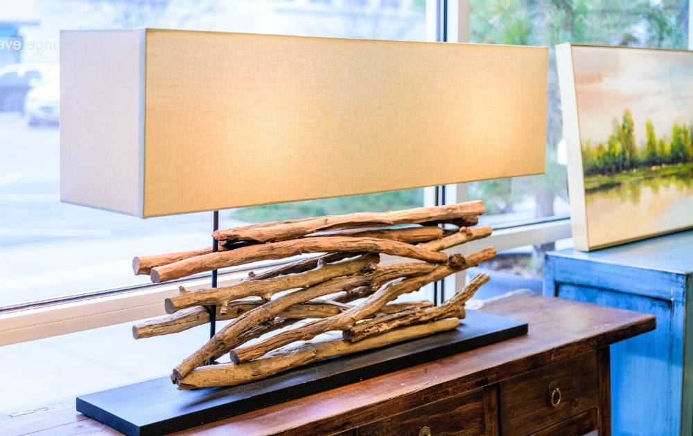 A close-up of a lit floor lamp with a gold stand and pull chain, white lampshade, and blurred framed artwork visible on a beige wall in the background.