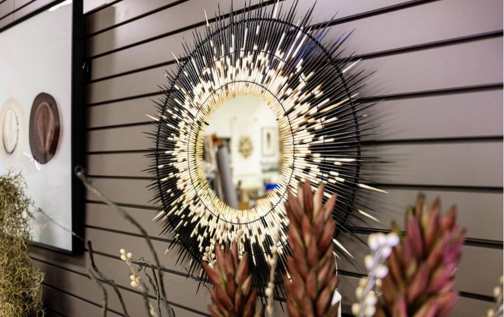 A round sunburst mirror with black and gold spokes hangs on a brown slatted wall, reflecting part of a room. Decorative plants and hats are displayed nearby.