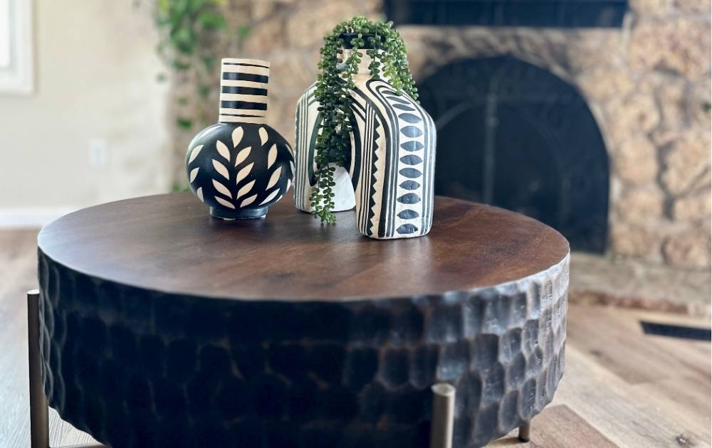 Two patterned black-and-white vases, one round and one rectangular with trailing greenery, sit on a dark wooden coffee table in front of a stone fireplace.