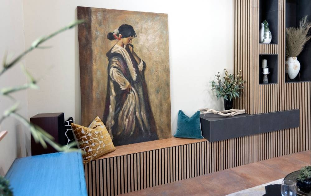 A large painting of a woman in a long dress leans against a modern wooden wall with built-in shelves. Cushions and decorative plants accent the minimalist, cozy sitting area.