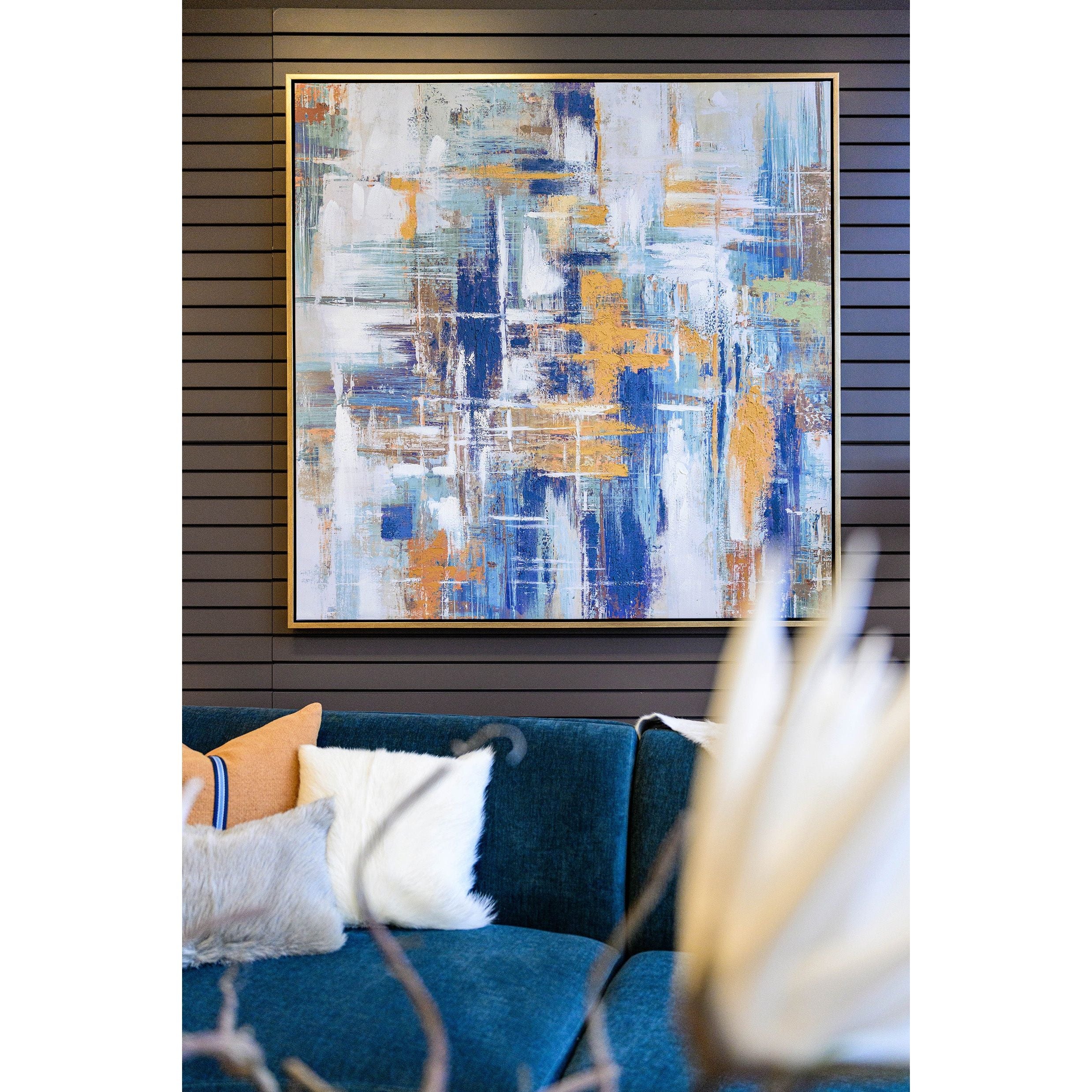 The Multicolored Abstract Canvas Wall Art, with bold blue, white, and orange brushstrokes, is elegantly framed on a dark paneled wall. Below it sits a teal couch adorned with white and orange cushions for contrast. A blurred foreground object adds depth to this contemporary space.