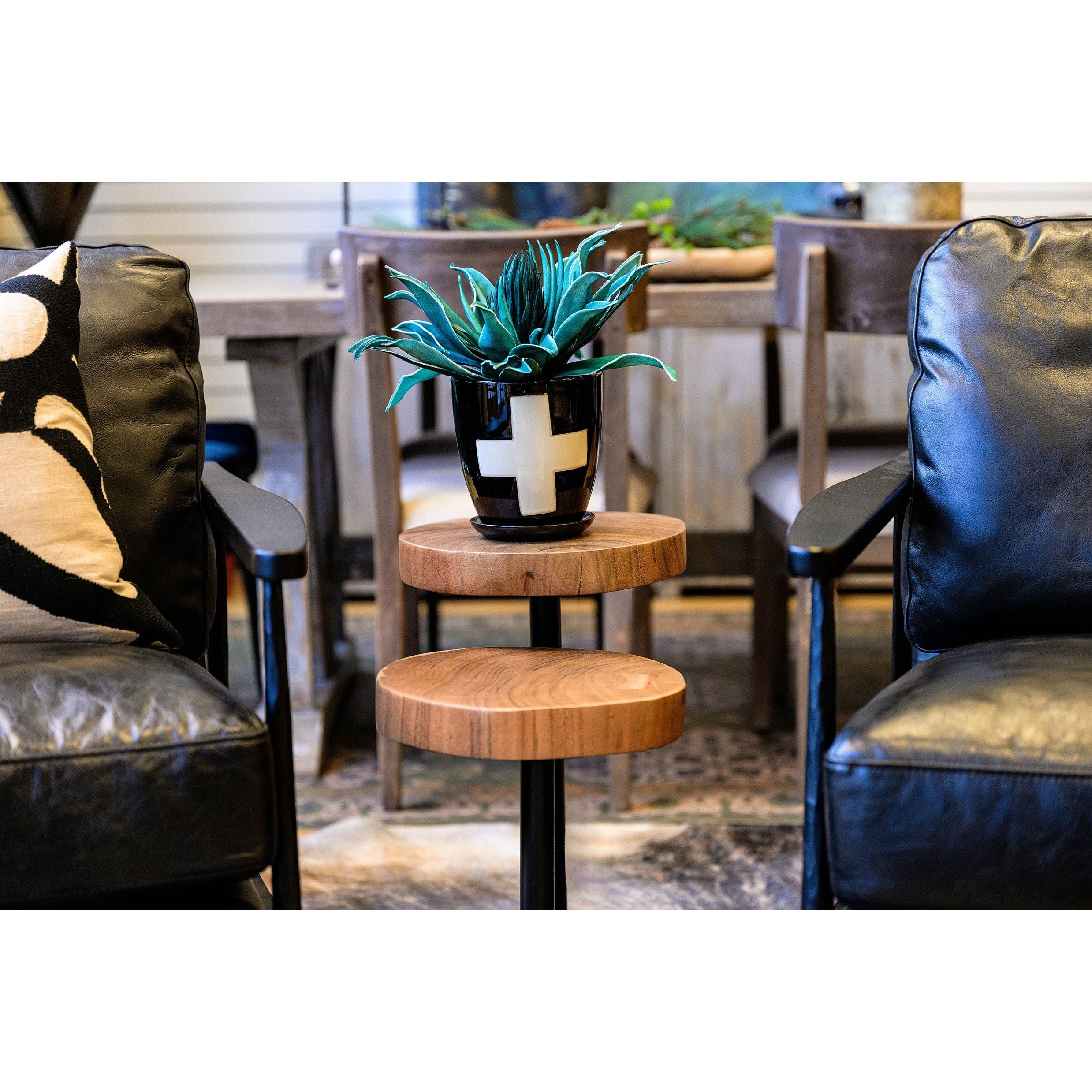 A set of two Nesting Martini Side Tables, crafted from solid acacia wood with three staggered tiers, stands between black leather chairs. A black pot with a white plus sign and a green succulent tops one table; unique dining pieces blur behind.