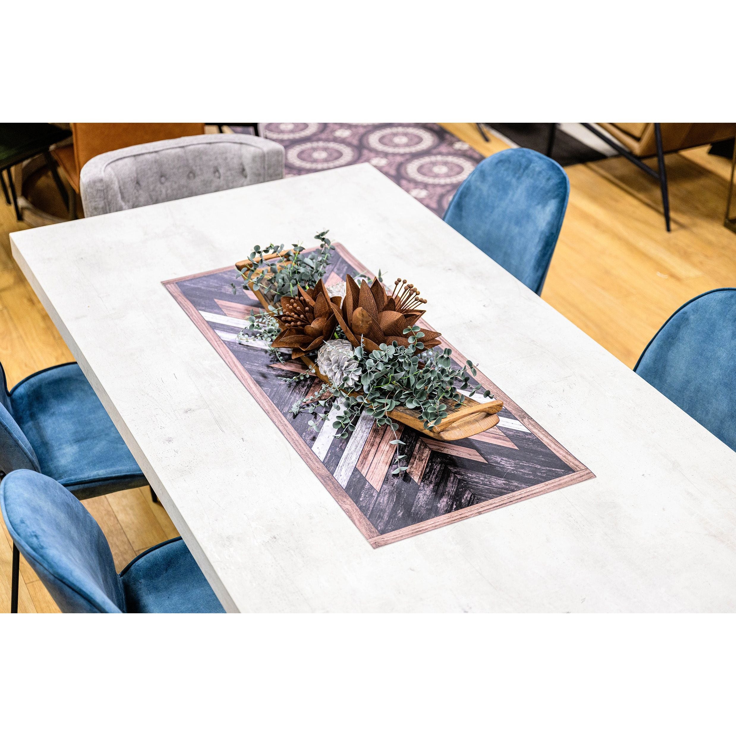 The Drew Dining Table, crafted from FSC Certified Reclaimed Wood, sits on a wooden floor and features a geometric-patterned runner with a eucalyptus and dried flower centerpiece. It is surrounded by six blue velvet chairs and one gray chair, with part of a patterned rug visible below.