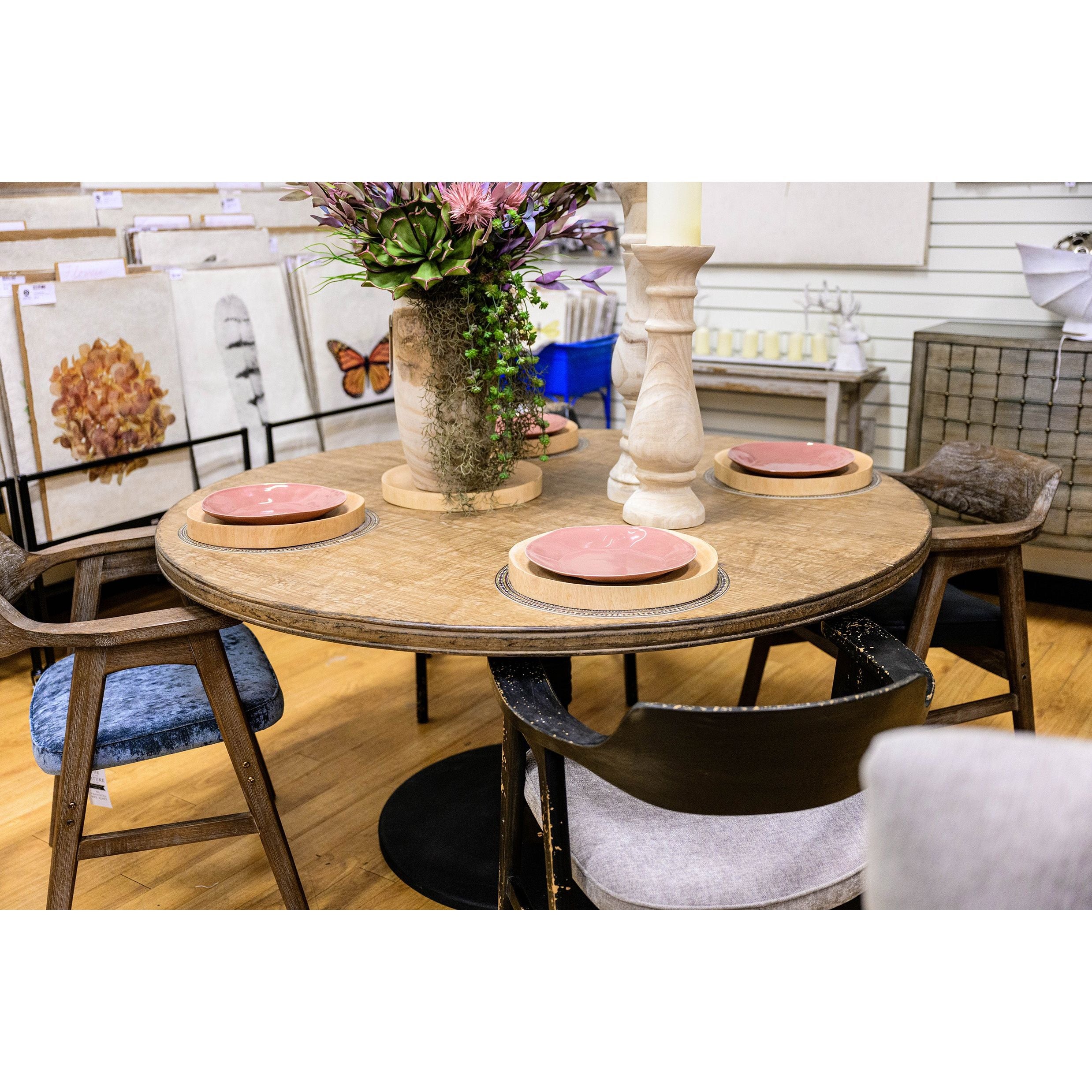 A round Sadie Dining Table made from reclaimed pine wood stands elegantly with four place settings, each featuring a pink plate on a woven placemat. The table is decorated with a tall vase of flowers and a smaller vase. Wooden chairs with various cushion styles surround the table, framed artwork and decor in the background.