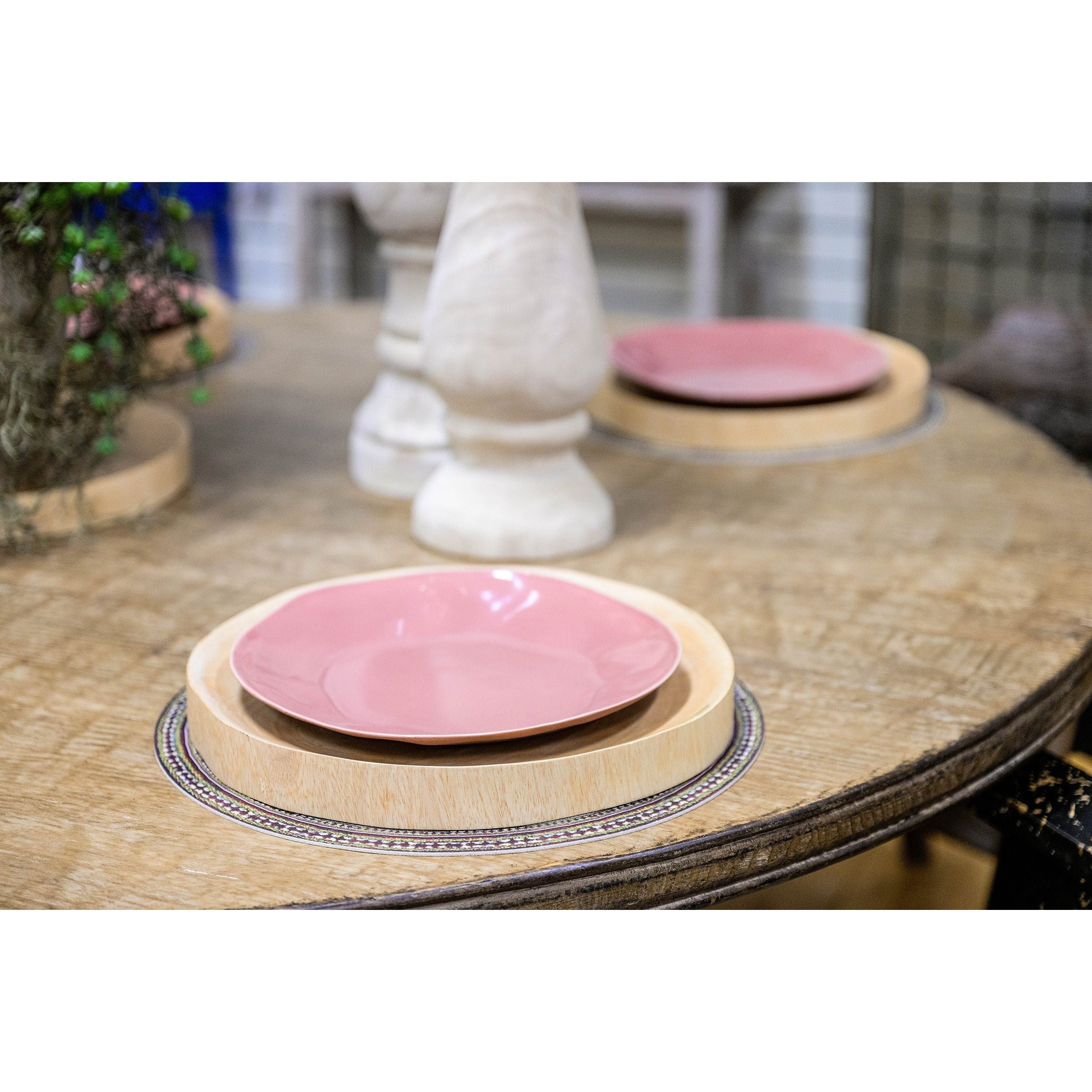 A round reclaimed pine wood table set for dining, featuring a pink plate placed on a light-colored circular tray. The table, known as the Sadie Dining Table, has additional trays with pink plates, a potted plant, and a tall decorative white candlestick holder. The hand-forged iron pedestal base adds elegance. The background includes blurred elements, suggesting an indoor setting.