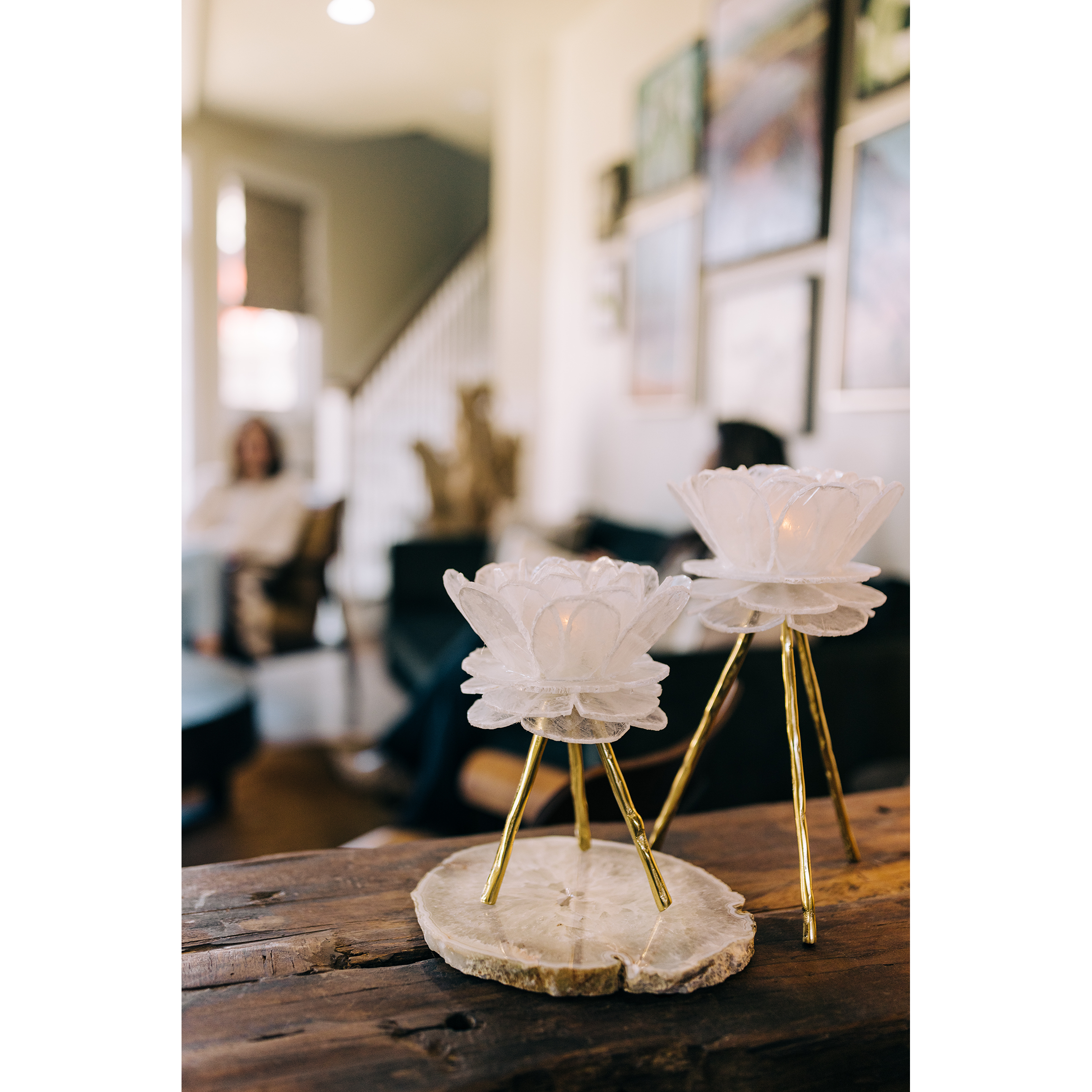 Two Selenite-Flower Candleholders, white and lotus-shaped with gold legs, stand on a textured wooden surface. The softly blurred background adds an ethereal glow to the cozy living room, featuring wall art and a staircase.