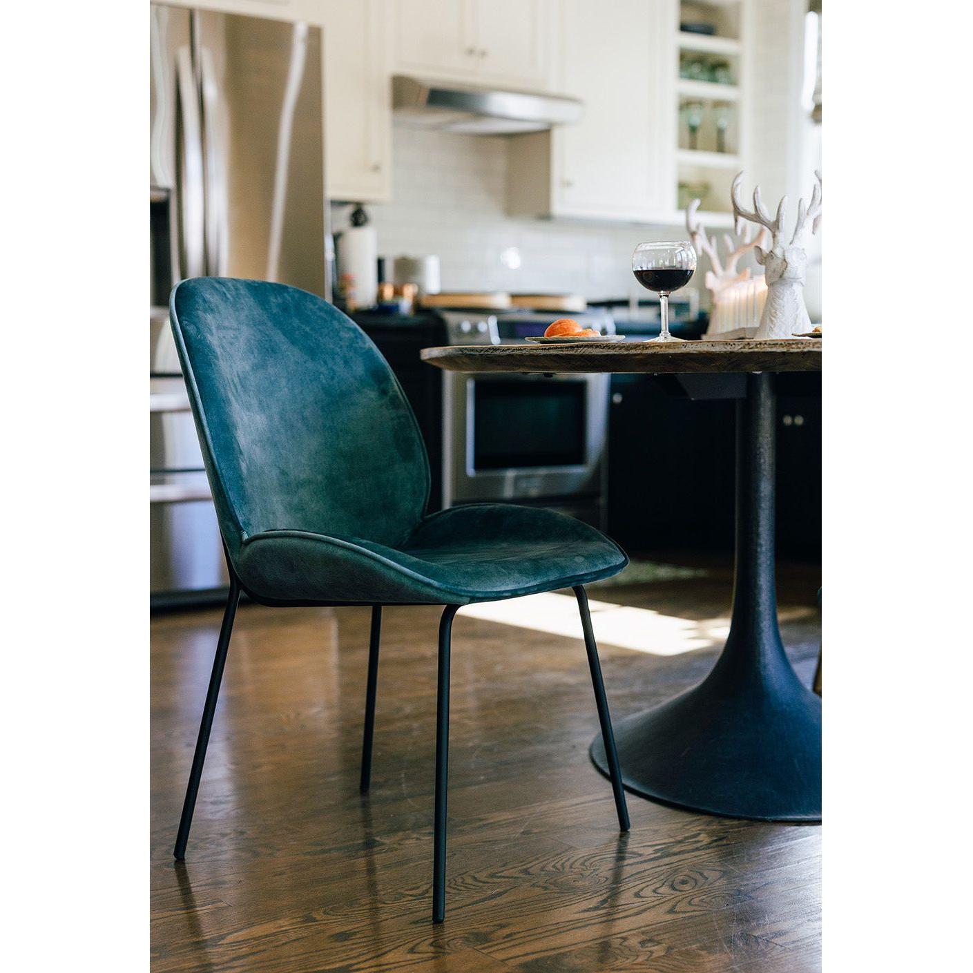 A modern kitchen features a King Dining Chair, Smoked Turquoise with metal legs beside a round table. On the table are a glass of red wine, a white coral decoration, and an orange. The kitchen boasts stainless steel appliances, white cabinets, and wooden flooring. Shelves with dishes are visible in the background.