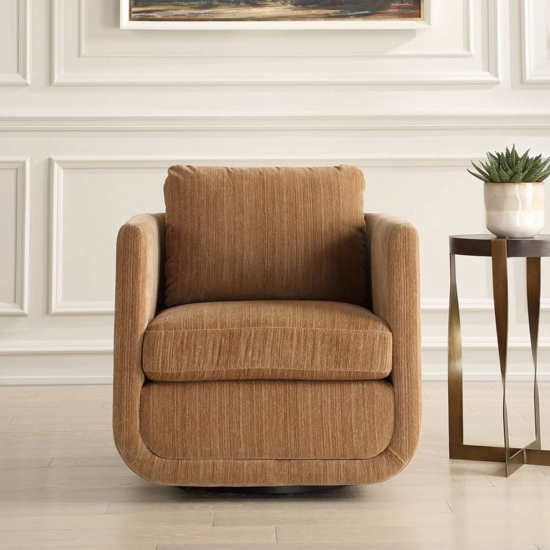 A Solana Swivel Accent Chair in Ginger with sculptural curves and brown textured chenille sits on a light wood floor. To the right, a gold side table holds a white planter with a green succulent. White paneled walls and art complete the scene.
