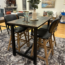 The Analee Counter Table features reclaimed pine and a hand-forged iron base, surrounded by four tall chairs with wooden legs and black seats. Decor is completed with two glasses, small saucers, and a white vase with green foliage on a patterned rug.