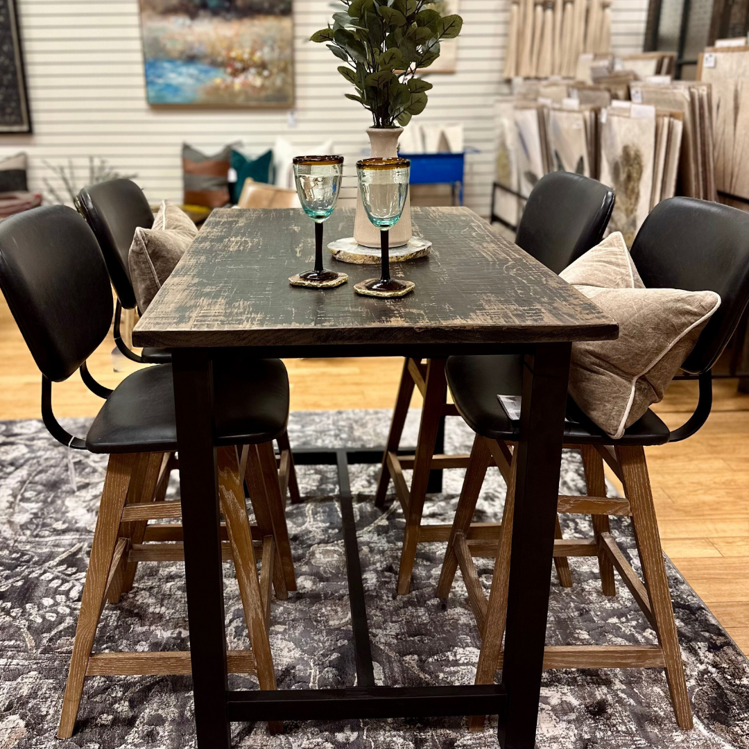 The Analee Counter Table with a hand-forged iron base is styled with blue glasses and coasters. Surrounded by four black chairs with wood legs and beige pillows, it sits on a patterned rug amid art decor in the home decor store.