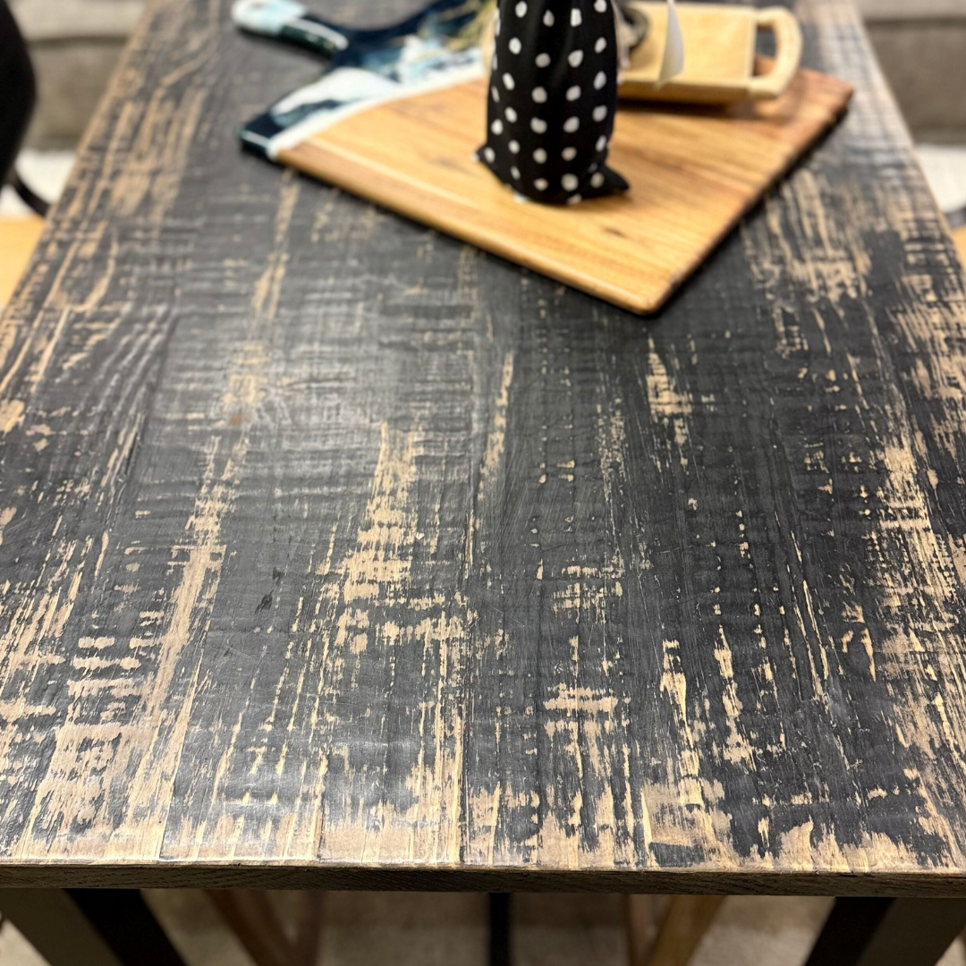 A close-up of the Analee Counter Table, crafted from reclaimed pine with a distressed black finish that reveals natural wood beneath. On the table, a wooden tray holds a black-and-white polka dot object and small items, fabric blurred in the background.