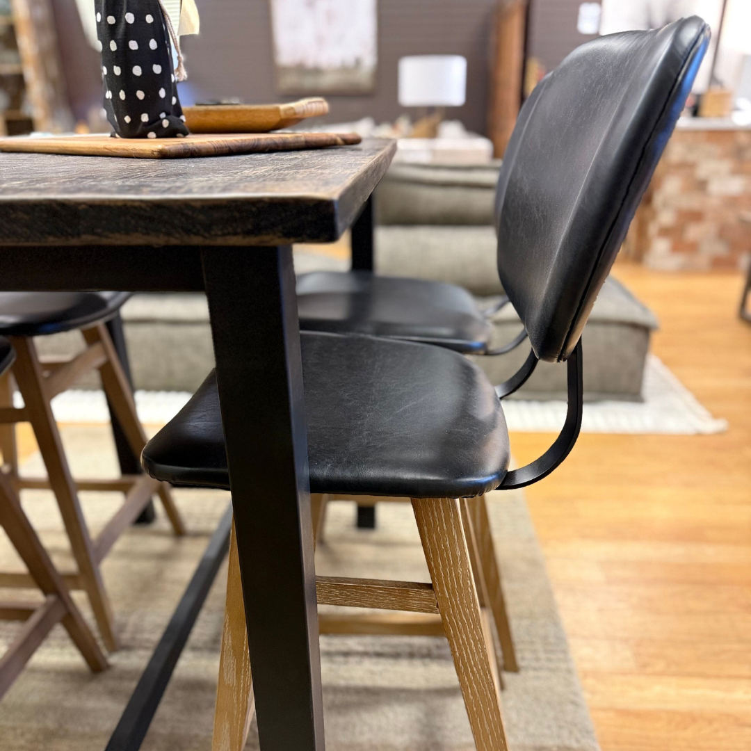 Two modern bar stools with black leather seats, wooden legs, and iron bases sit at an Analee Counter Table on a beige rug in a warmly lit room with wooden flooring.