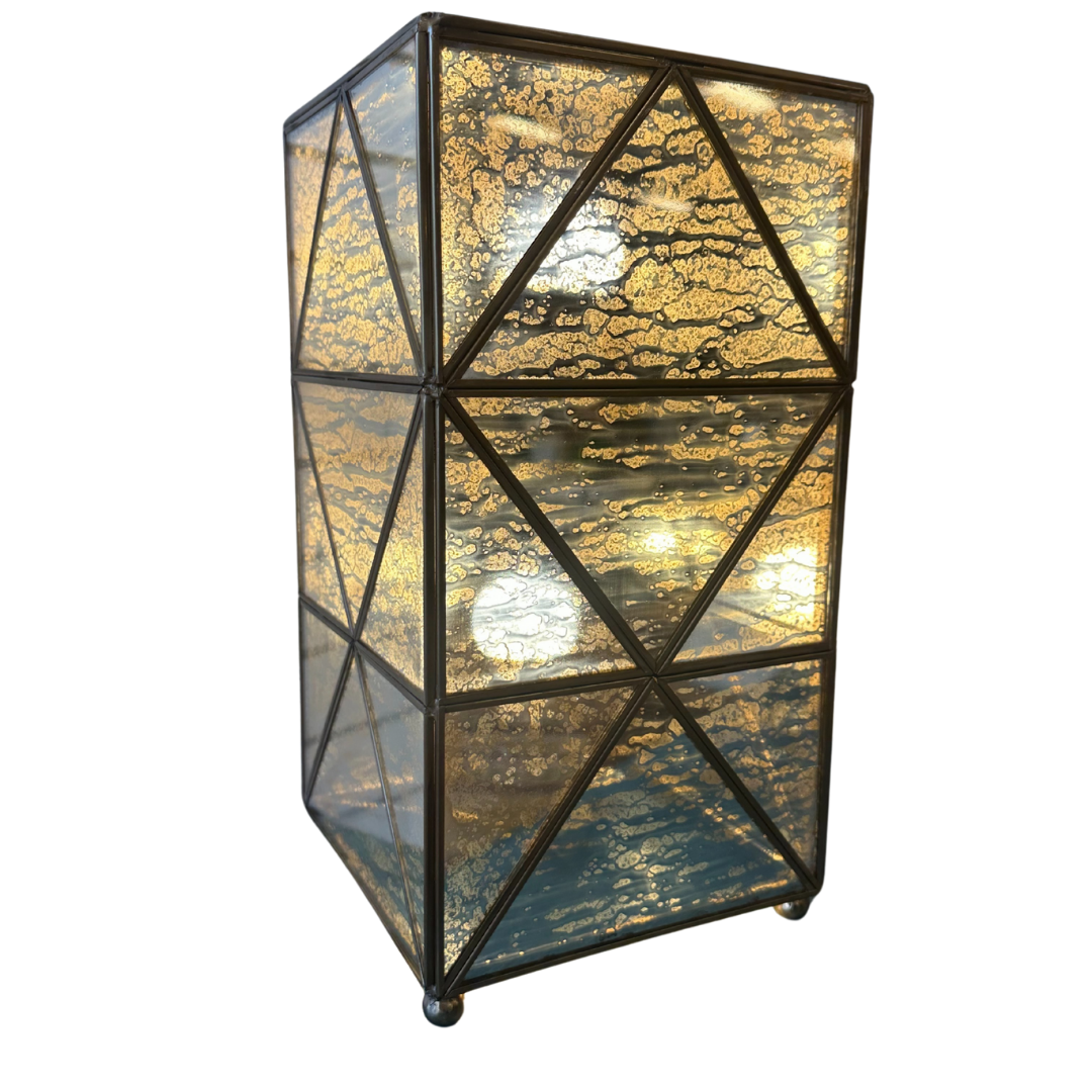 The Belvoir Gold-Trimmed Glass Lamp features a black metal frame and triangular mirrored glass panels with a marbled pattern in blue and gold. It emits a warm glow, standing on four small spherical feet for vintage glamour.