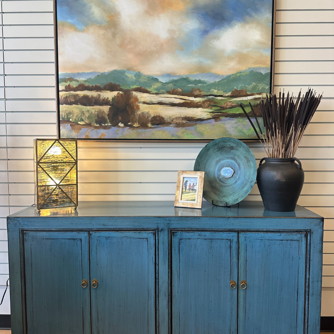 A blue sideboard with four paneled doors stands against a striped wall. Atop it are the Belvoir Gold-Trimmed Glass Lamp with a geometric design, a brass-framed picture, a green decorative plate, and a black vase with tall sticks. Above them hangs a large landscape painting of mountains and fields.