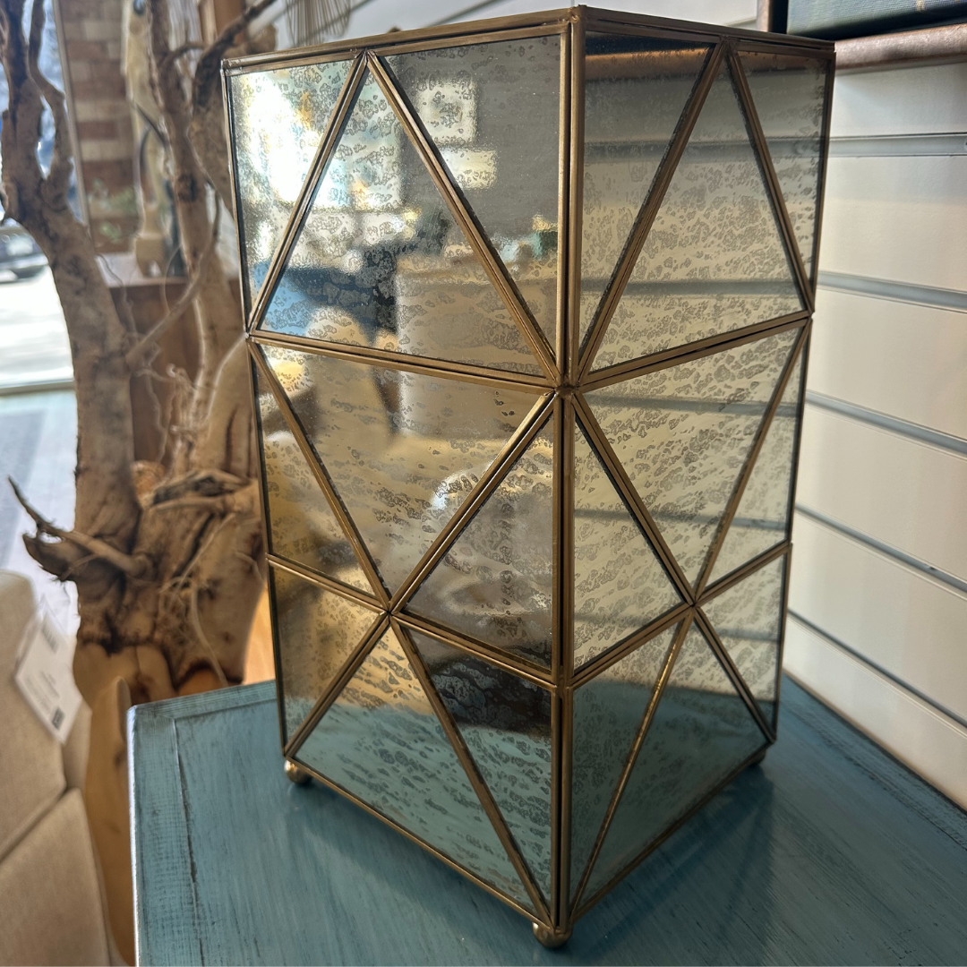 The Belvoir Gold-Trimmed Glass Lamp, with a brass frame and triangular panels, rests on a teal wooden table. The glass has an antique mottled finish. In the background, a blurred window display and tree branch decoration complement the elegant brass frame perfectly.