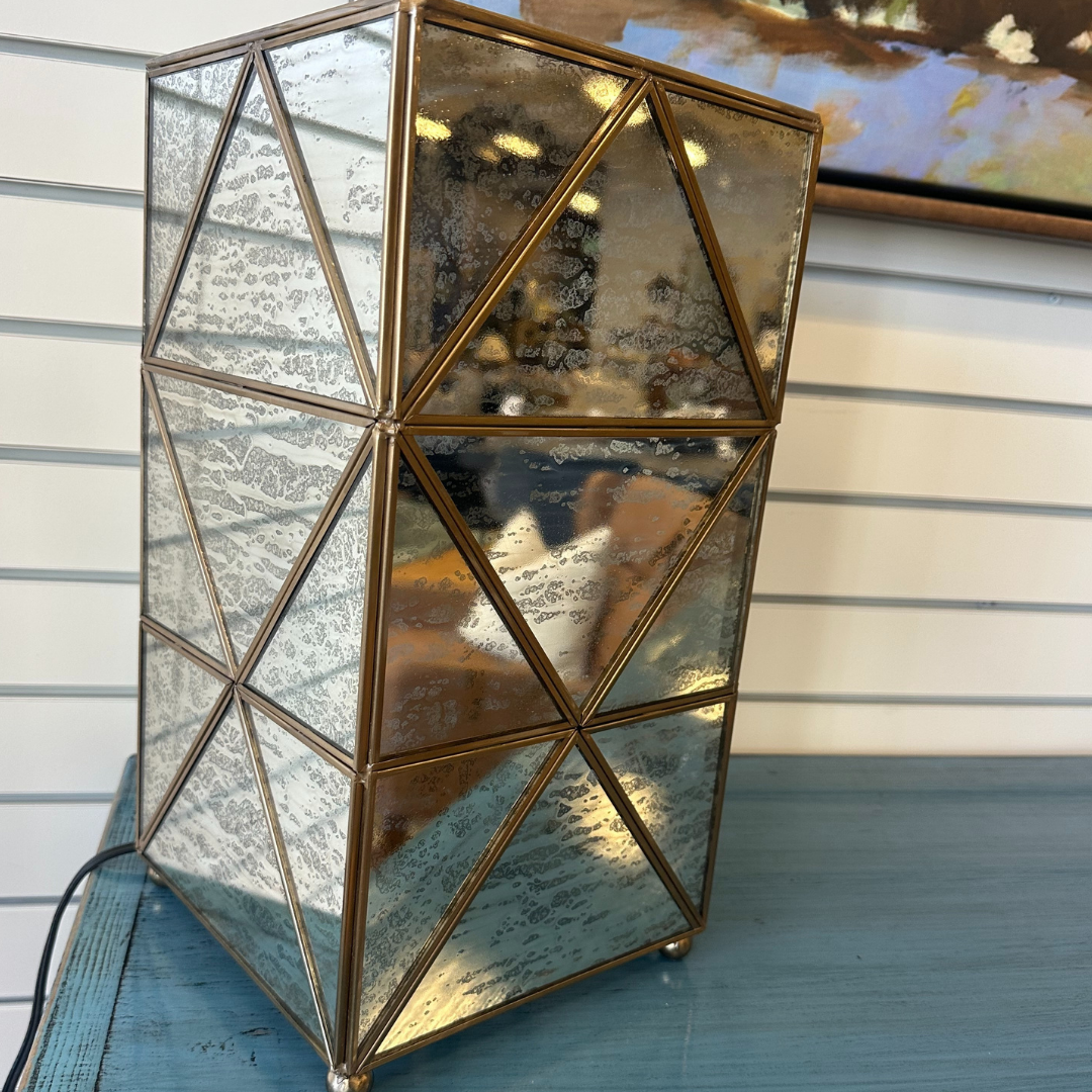 The Belvoir Gold-Trimmed Glass Lamp, featuring a geometric design with mirrored glass panels and a bronze frame, rests elegantly on a teal surface against a white, horizontally-striped wall, with a painting partially visible in the background adding color.