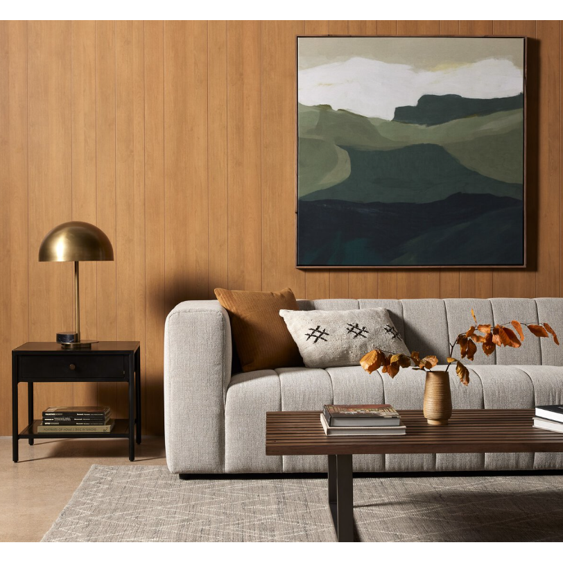 A modern living room features a light gray sofa adorned with a patterned pillow and brown cushion. In front, there's a wooden coffee table holding a vase of dried leaves. The Samuel Side Table, designed with black-finished iron legs, supports a brass lamp to the side. A large abstract painting highlighting green and white shapes hangs on the wood-paneled wall.