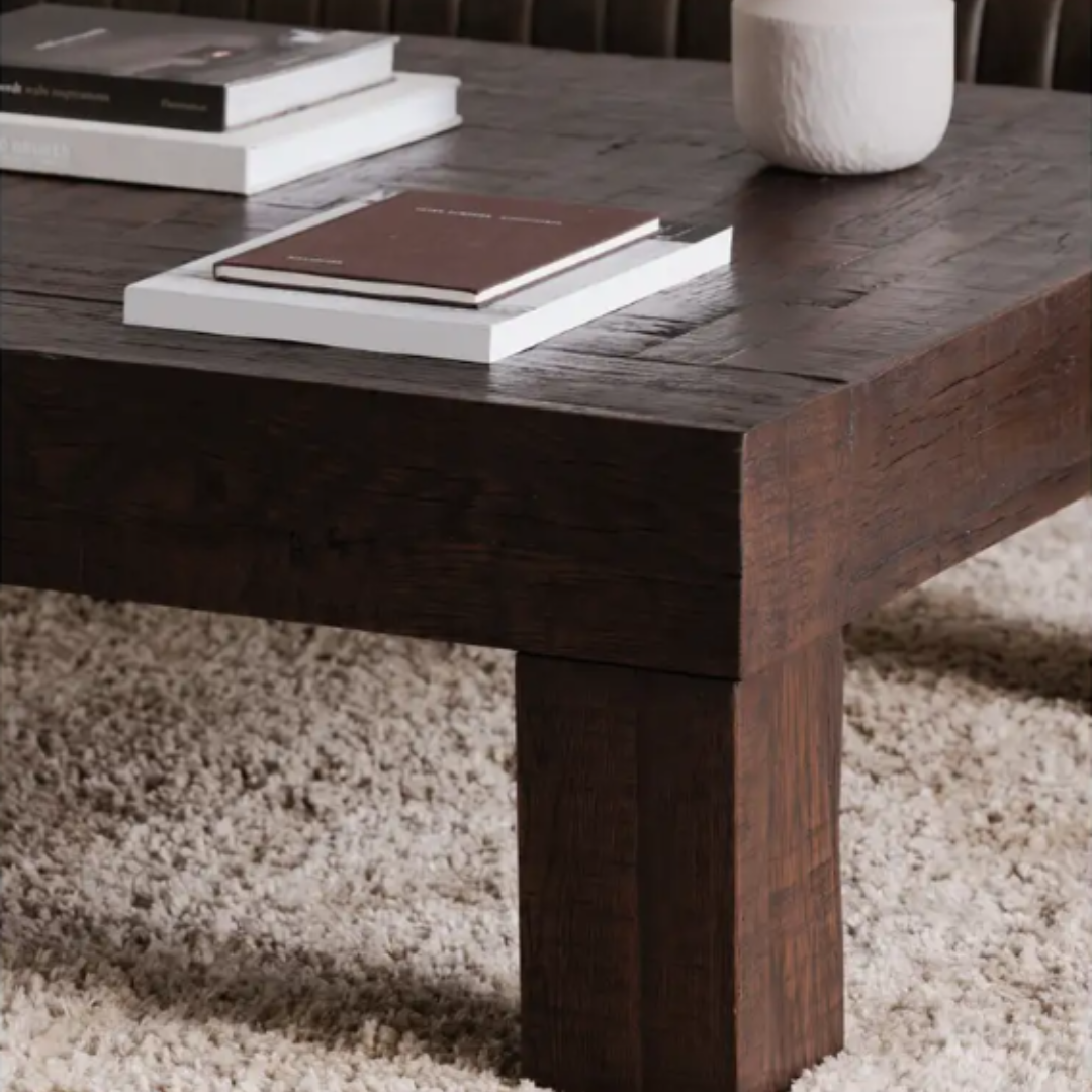 A close-up of the Block Coffee Table in rustic brown sits on a textured beige carpet. Two stacked books and a small white ceramic vase rest on its dark, natural wood surface with visible grain and sturdy legs, while the background features a hint of a brown sofa.