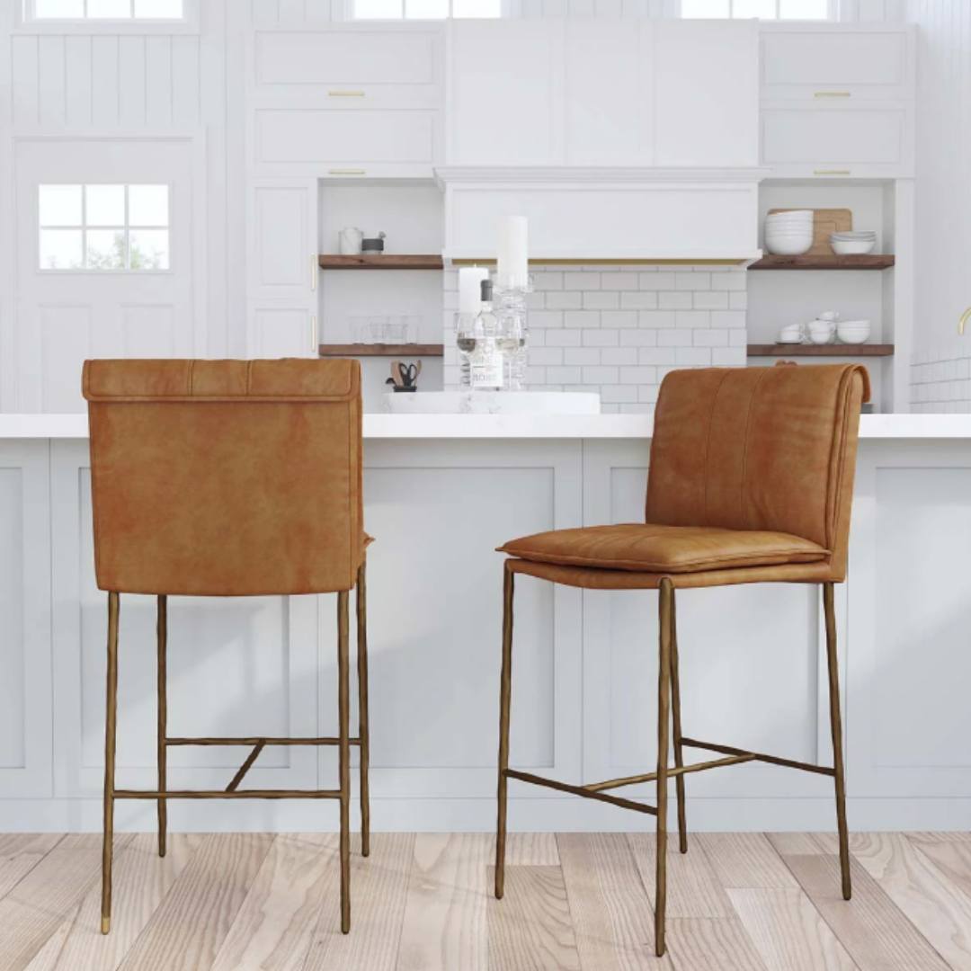 Two Calder Leather Counter Stools with sculptural metal frames stand before a white kitchen island. The space features white cabinets, subway tile backsplash, open shelves, and decor like candle holders and glassware on a light wood floor.