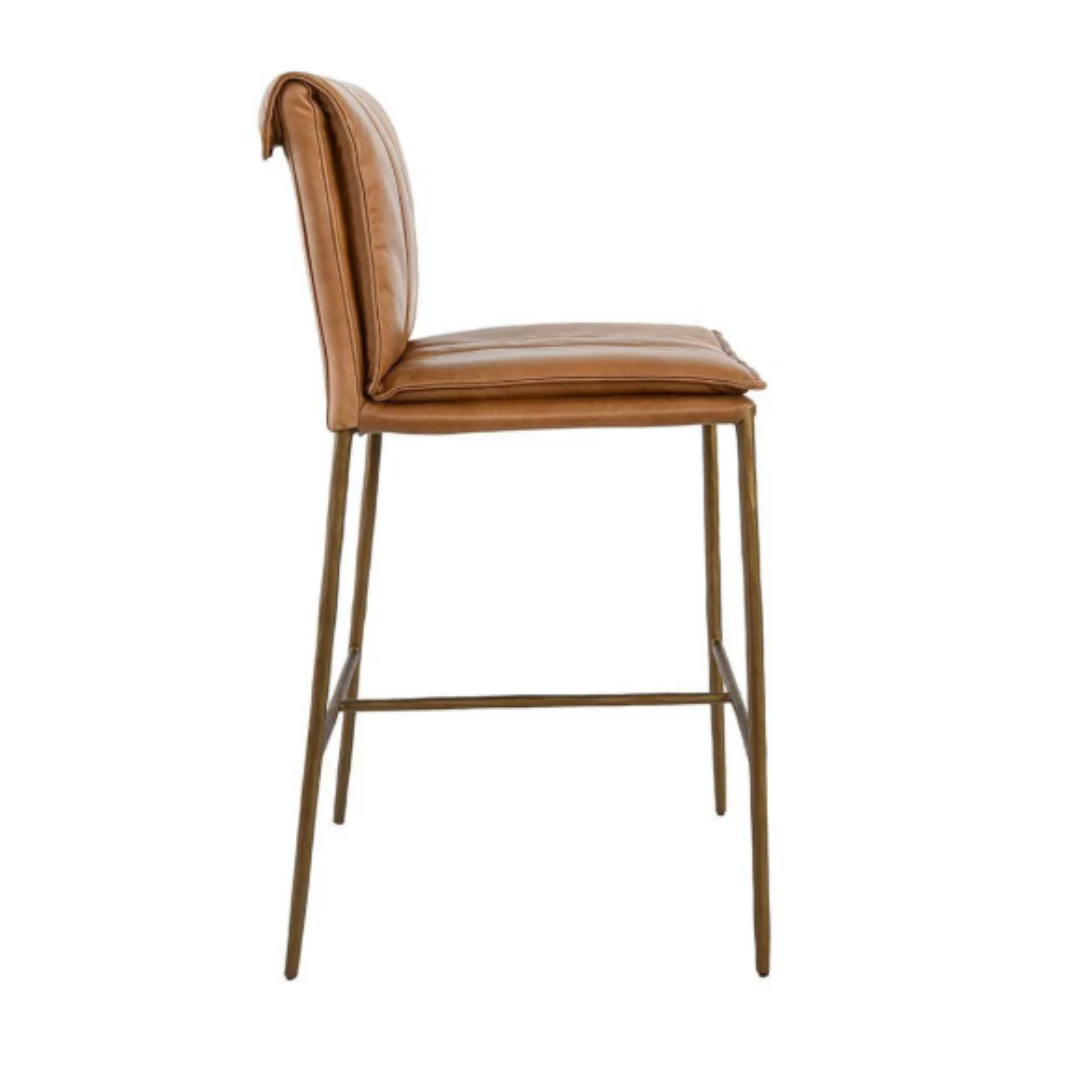 The Calder Leather Counter Stool showcases a sculptural metal frame with slender, gold-toned legs and light brown leather upholstery. A footrest links the front pair of legs, enhancing its sleek and minimalist design.