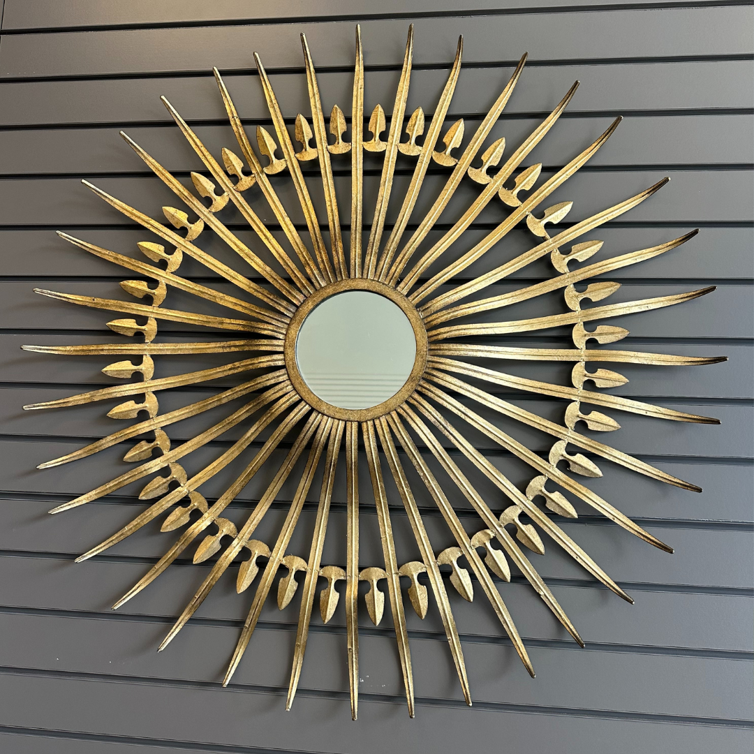 The Cali Gold Sunburst Wall Mirror adorns the panelled grey wall with timeless elegance, featuring radiating spikes and intricate cut-out designs near the center that exude vintage glamour resembling a sun with an ornate flair.