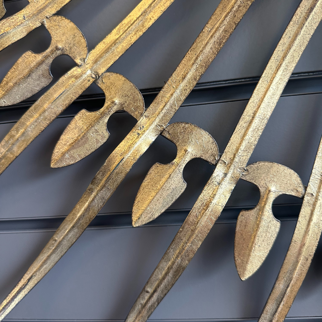 The Cali Gold Sunburst Wall Mirror features a close-up of metallic gold leaf-like shapes against a dark gray background, showcasing intricate vintage glamour with smooth curves and symmetrical forms that exude timeless elegance and craftsmanship.