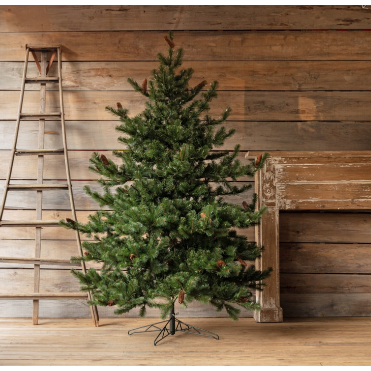 The Classic Christmas Tree (Pre-Lit) stands on a wooden floor between a weathered ladder and a rustic fireplace mantel, set against wood-paneled walls in a cozy, simply decorated room.