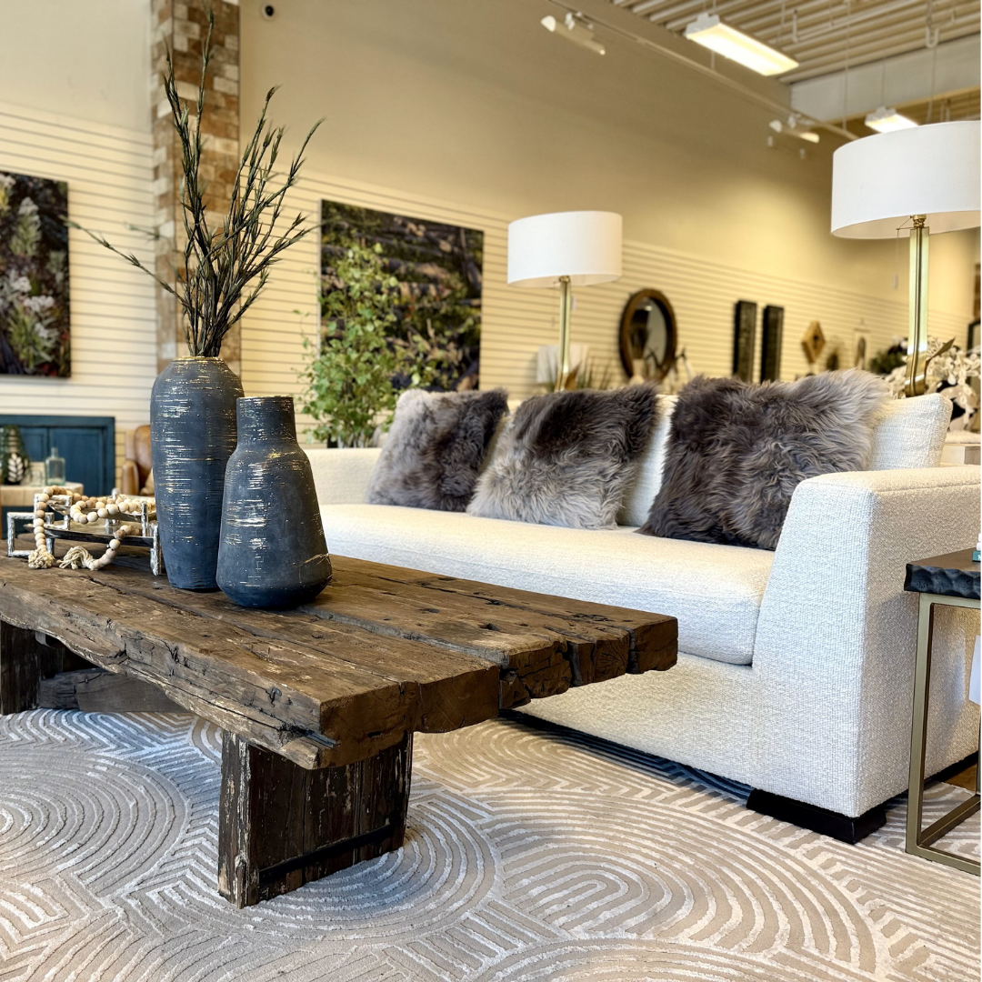 A modern living room showcases the Dresden Performance Fabric Customizable Sofa with shelter arms and gray fur pillows, paired with a rustic wooden coffee table, black vases, neutral wall art, floor lamps, and a patterned light gray rug for a cozy elegance.