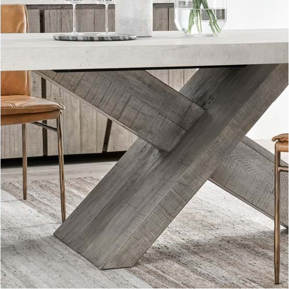The Drew Dining Table showcases a sustainably designed white concrete laminate top and textured gray wooden X-shaped legs, paired with brown leather chairs featuring metal legs. A neutral-toned rug lies beneath, and a glass vase with greenery adorns the background.