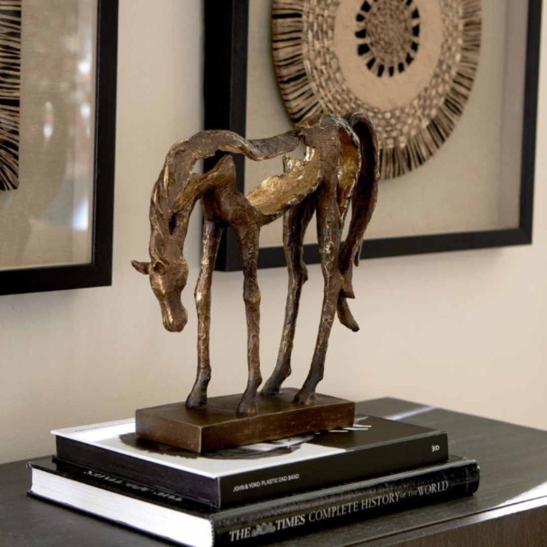 The Eclipse Horse Sculpture, elegantly slim in antiqued bronze, sits atop two books on a beige surface. Its minimalist design exudes timeless elegance, while three framed artworks with intricate geometric designs decorate the pale wall behind it.