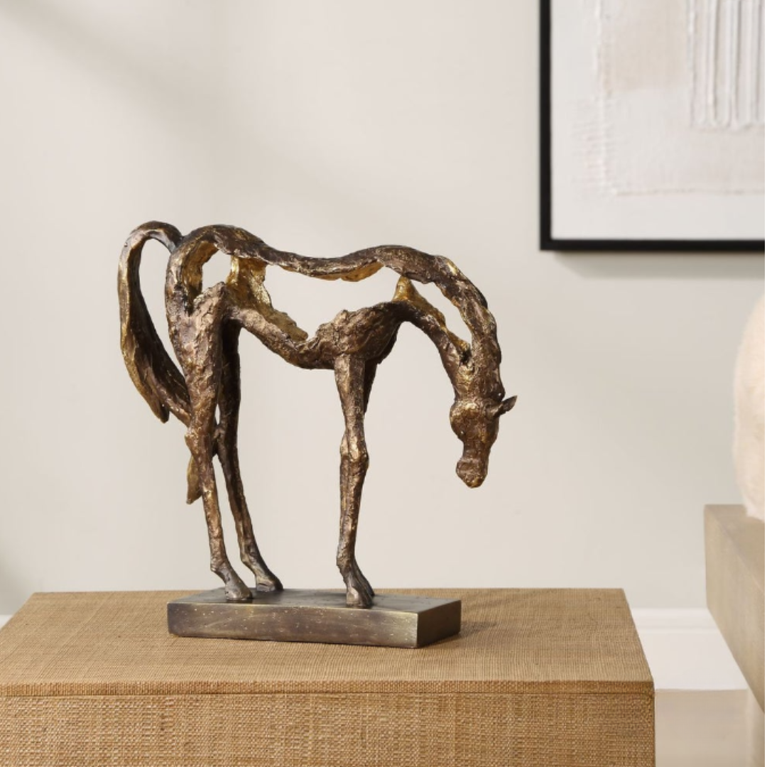 The Eclipse Horse Sculpture, in antiqued bronze with a textured abstract design, elegantly stands on a wooden surface. Its minimalist form and gracefully lowered head add timeless elegance against a framed artwork on the light-colored wall.