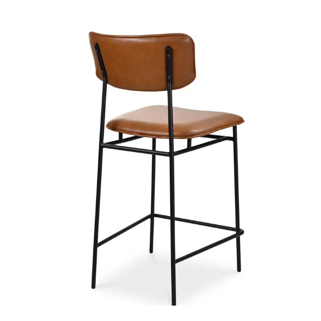 The Elara Leather Counter Stool boasts caramel-hued faux leather upholstery on the seat and backrest. Its black metal frame with four legs and footrests exemplifies modern minimalism, while the angled view highlights a sleek profile that effortlessly blends modern and industrial styles.