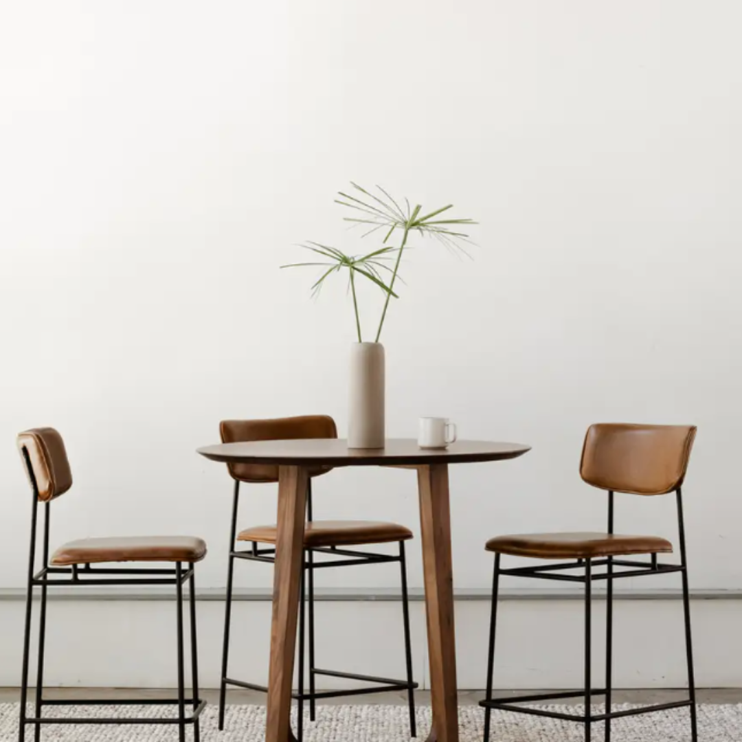 A modern minimalist dining setup showcases a round wooden table with a light brown finish, paired with three Elara Leather Counter Stools featuring caramel upholstery and metal frames. On the table, a tall vase holding sparse greenery and a white mug complement the plain white wall backdrop.