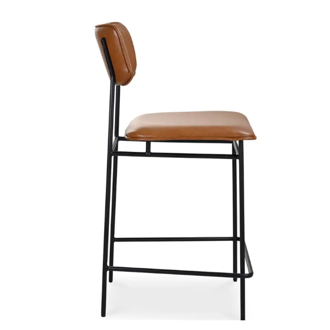 The Elara Leather Counter Stool boasts a caramel leather seat and backrest, epitomizing modern minimalism. Its sleek black metal frame includes a slightly curved back and an integrated footrest, adding to its simple yet sophisticated appeal.