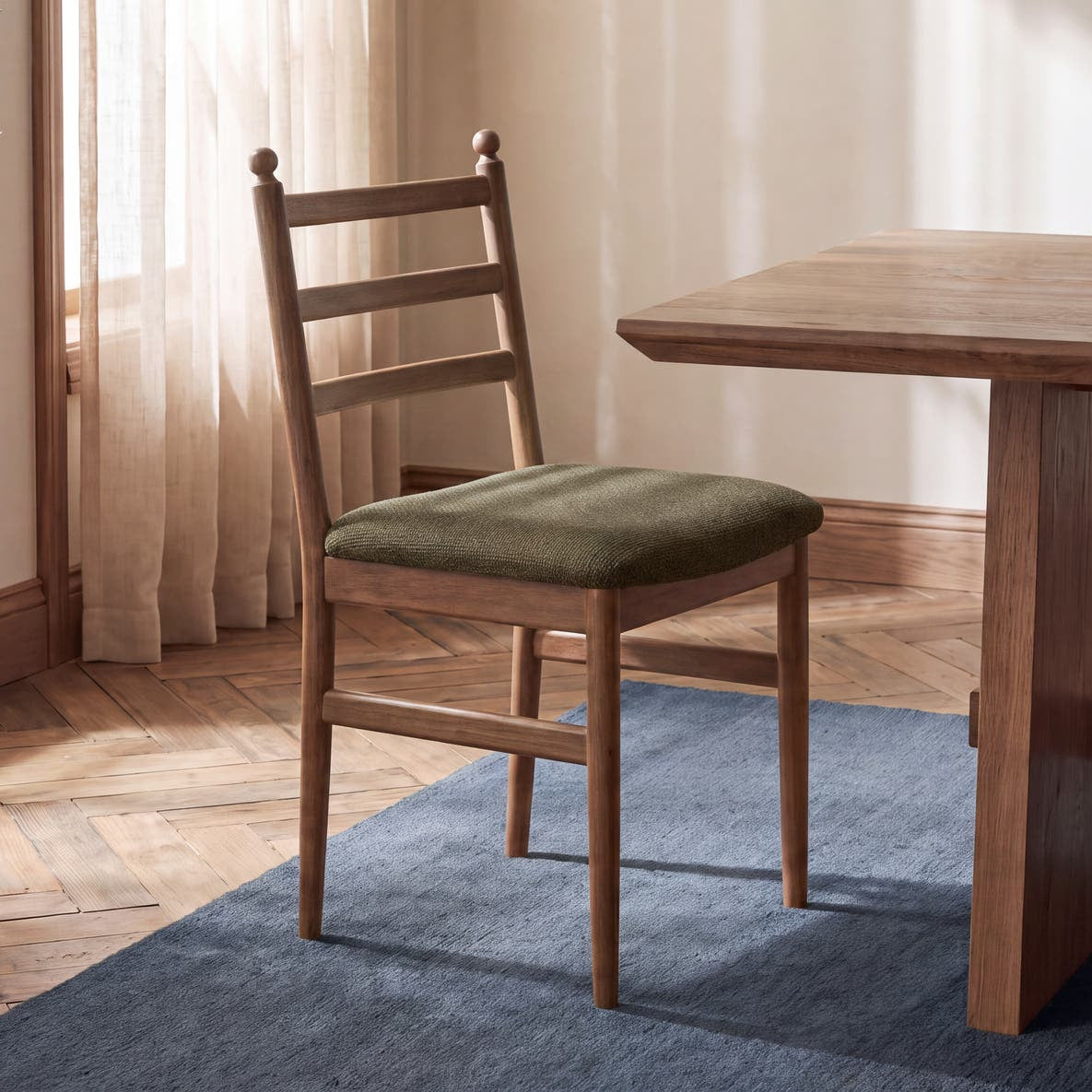 A wooden dining chair with a green upholstered seat cushion sits beside a matching wooden table in a warm, sunlit room with a blue rug, wood floor, and a potted plant.